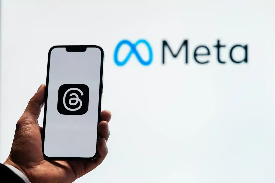 A hand holding a smartphone displaying the Threads app logo, with the Meta logo visible in the background. The image represents the Threads app as part of Meta's branding ecosystem, now that advertising on Threads is becoming available.